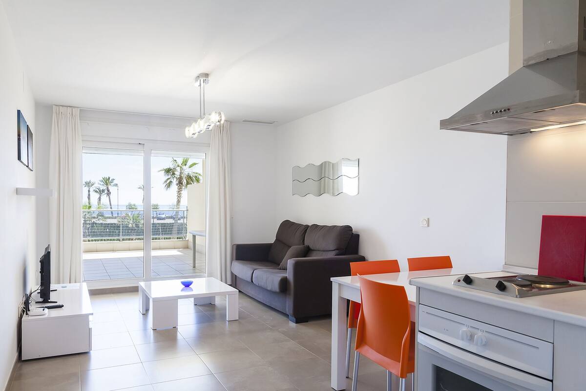 A modern kitchen with a gas stove, a dining area with orange chairs, a sofa, and a large window with a view of palm trees.