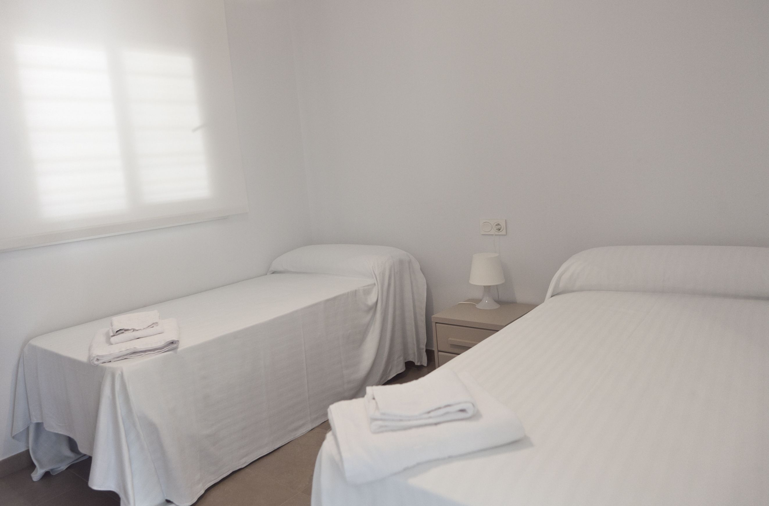 Una habitación con dos camas, una mesita de noche con lámpara y una ventana con persianas.