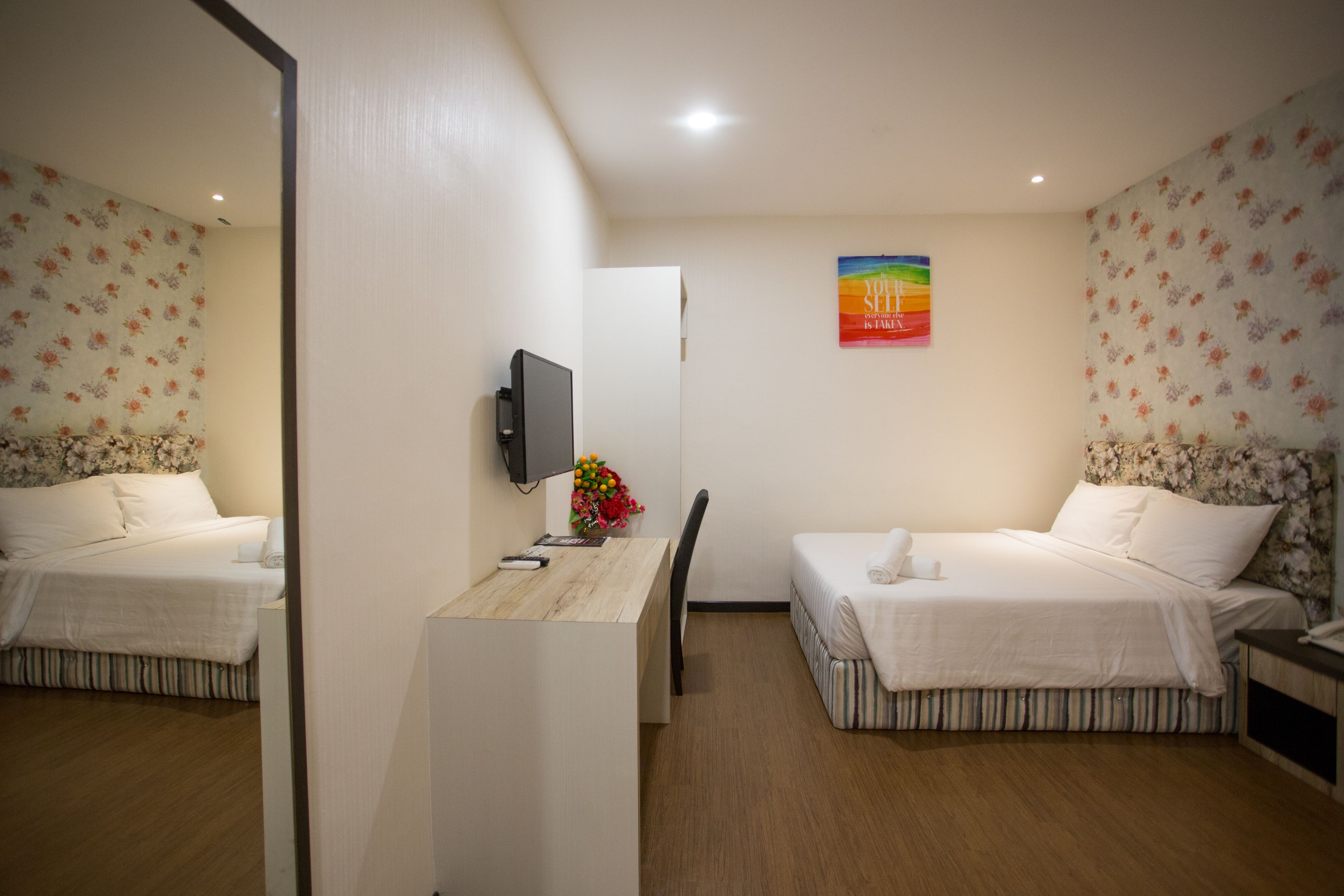 A hotel room with two beds, a desk, a chair, a TV, and a floral wallpaper.