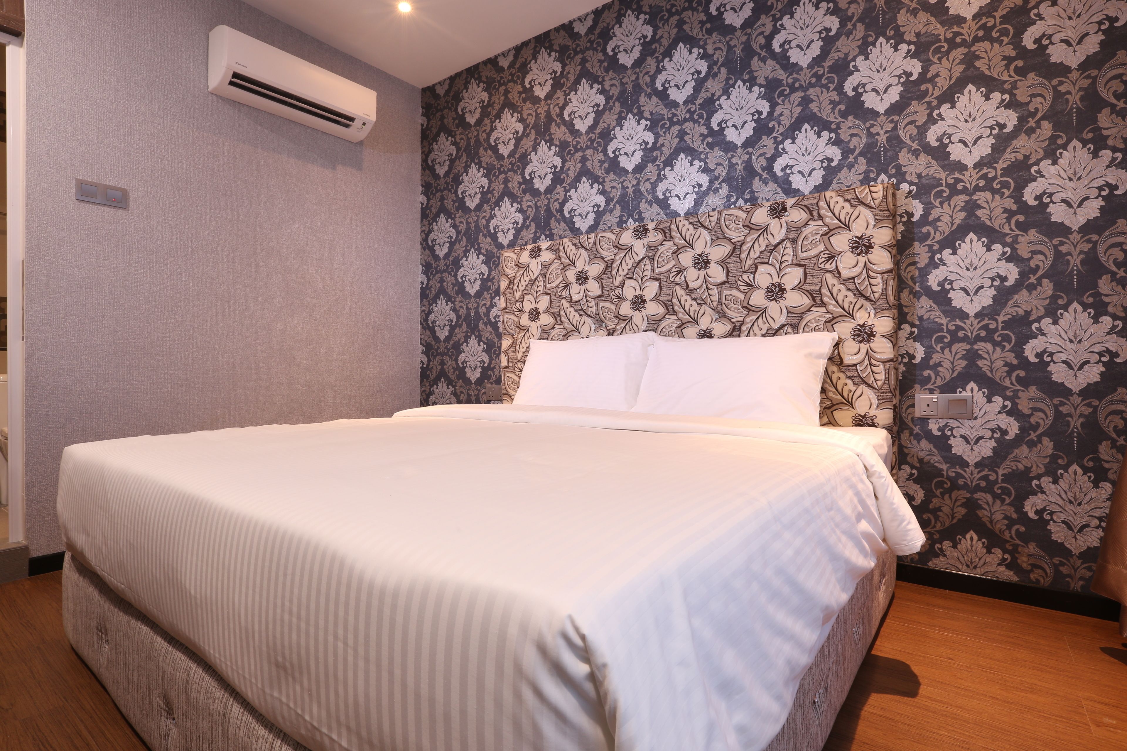 A neatly made bed with a white comforter and pillows, a patterned wallpaper, and a wall-mounted air conditioner.