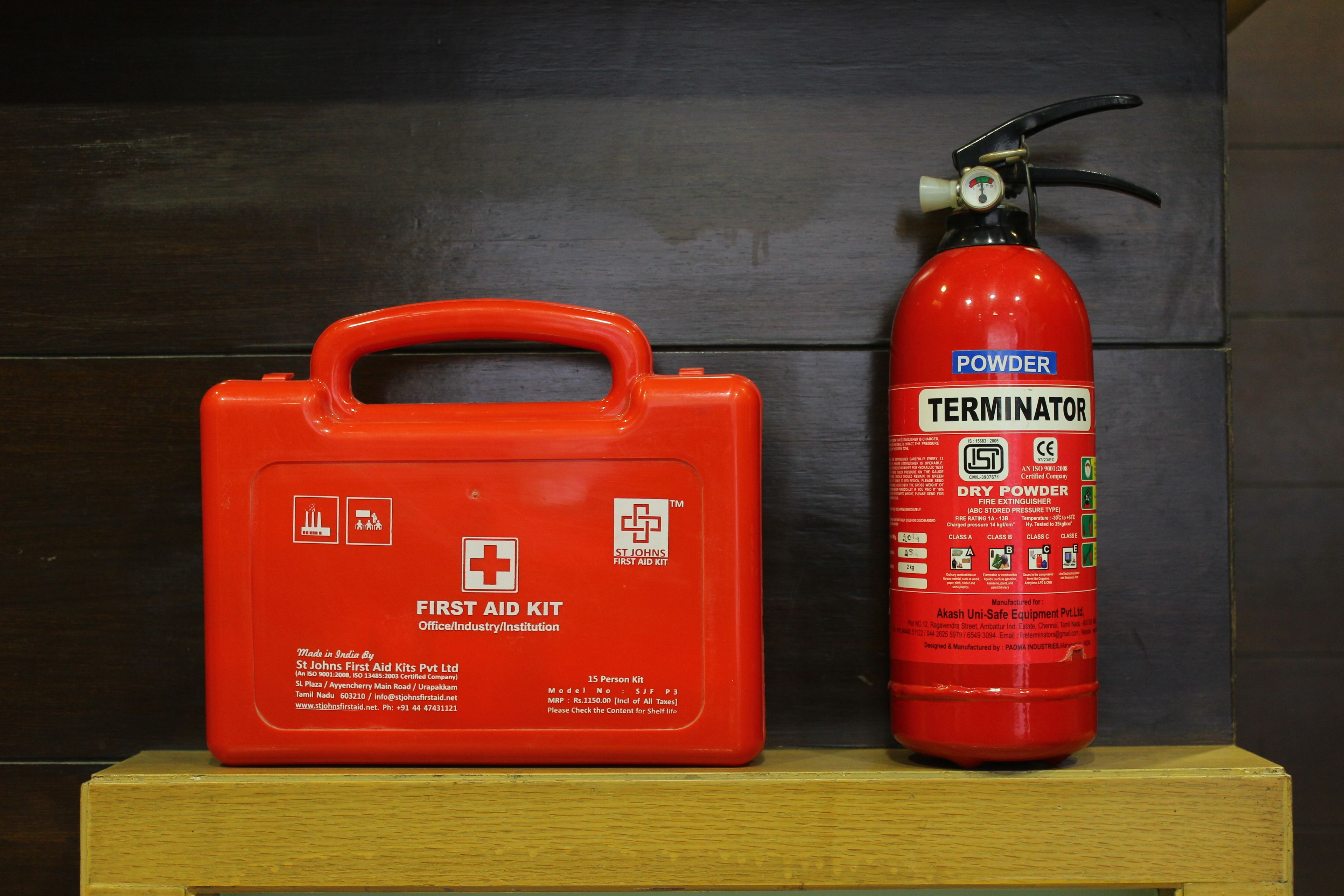 A red fire extinguisher and a red first aid kit on a wooden shelf against a dark background.
