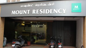 L’entrée de Mount Residency est signalée par une enseigne en anglais et dans une autre langue. On y voit deux scooters garés à l’extérieur et une chaise près de l’entrée.