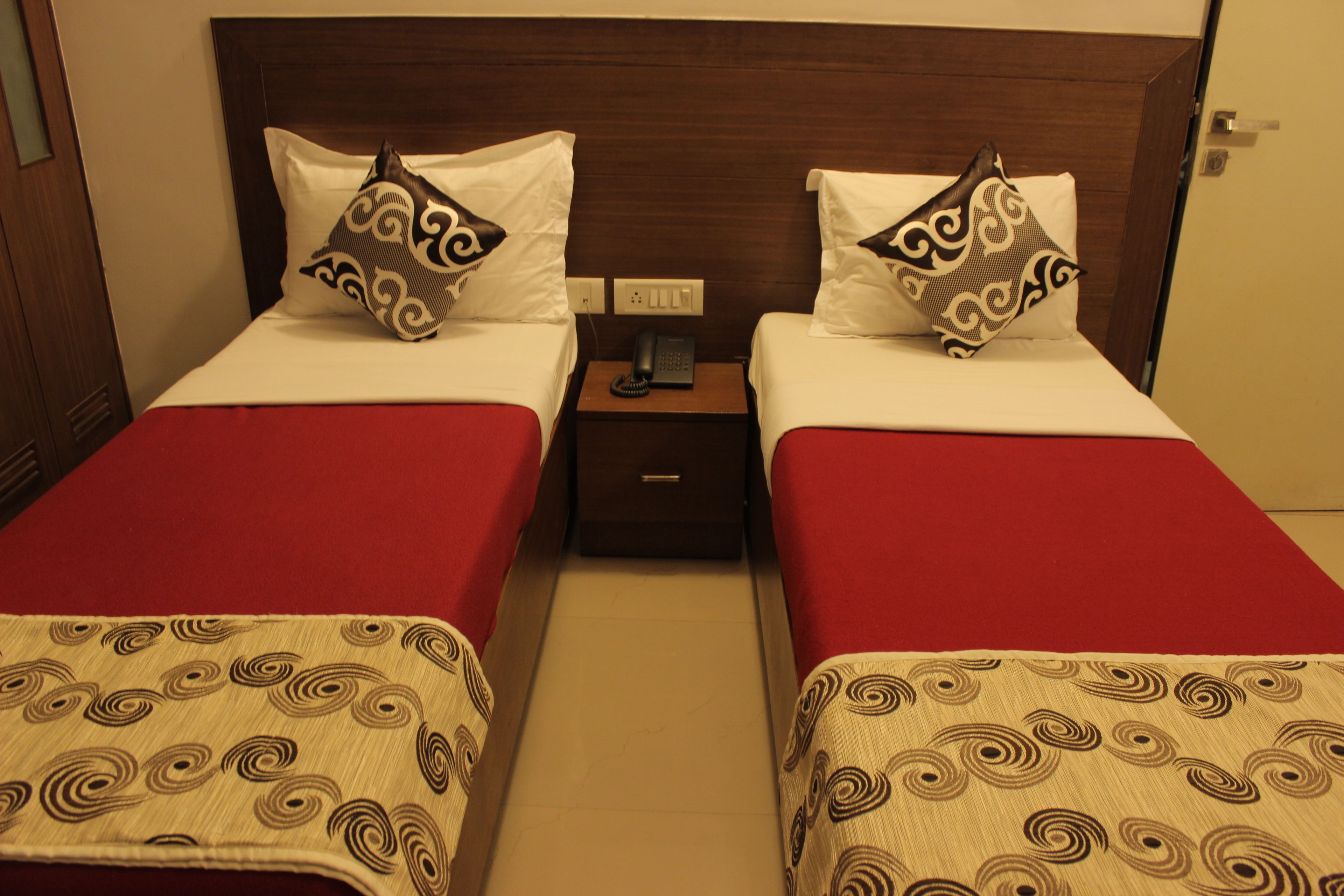 A hotel room with two beds, each with a red bedspread and a patterned throw. There is a wooden headboard, a bedside table with a phone, and a nightstand with a lamp.