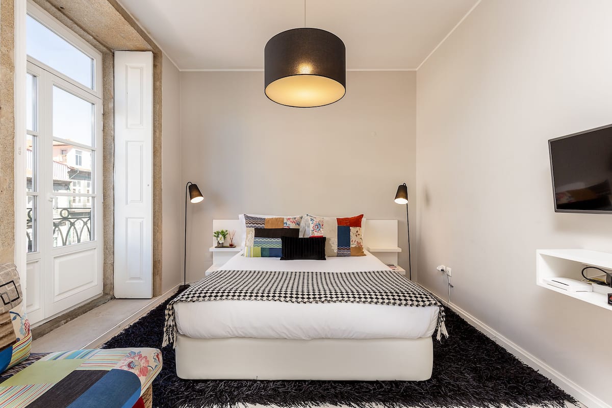 A modern bedroom with a large bed, a black and white patterned bedspread, two bedside lamps, a television on the wall, and a window with a view of buildings.
