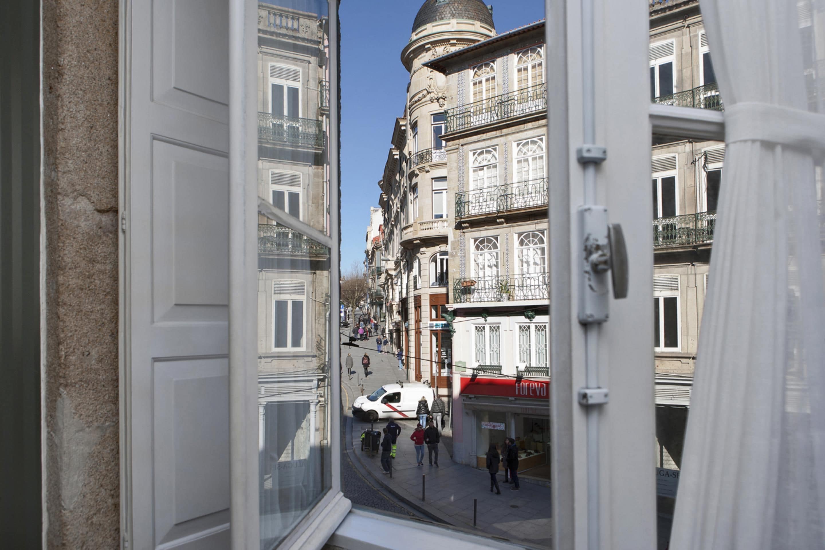A view through an open window of a street with buildings, a white van, and pedestrians.