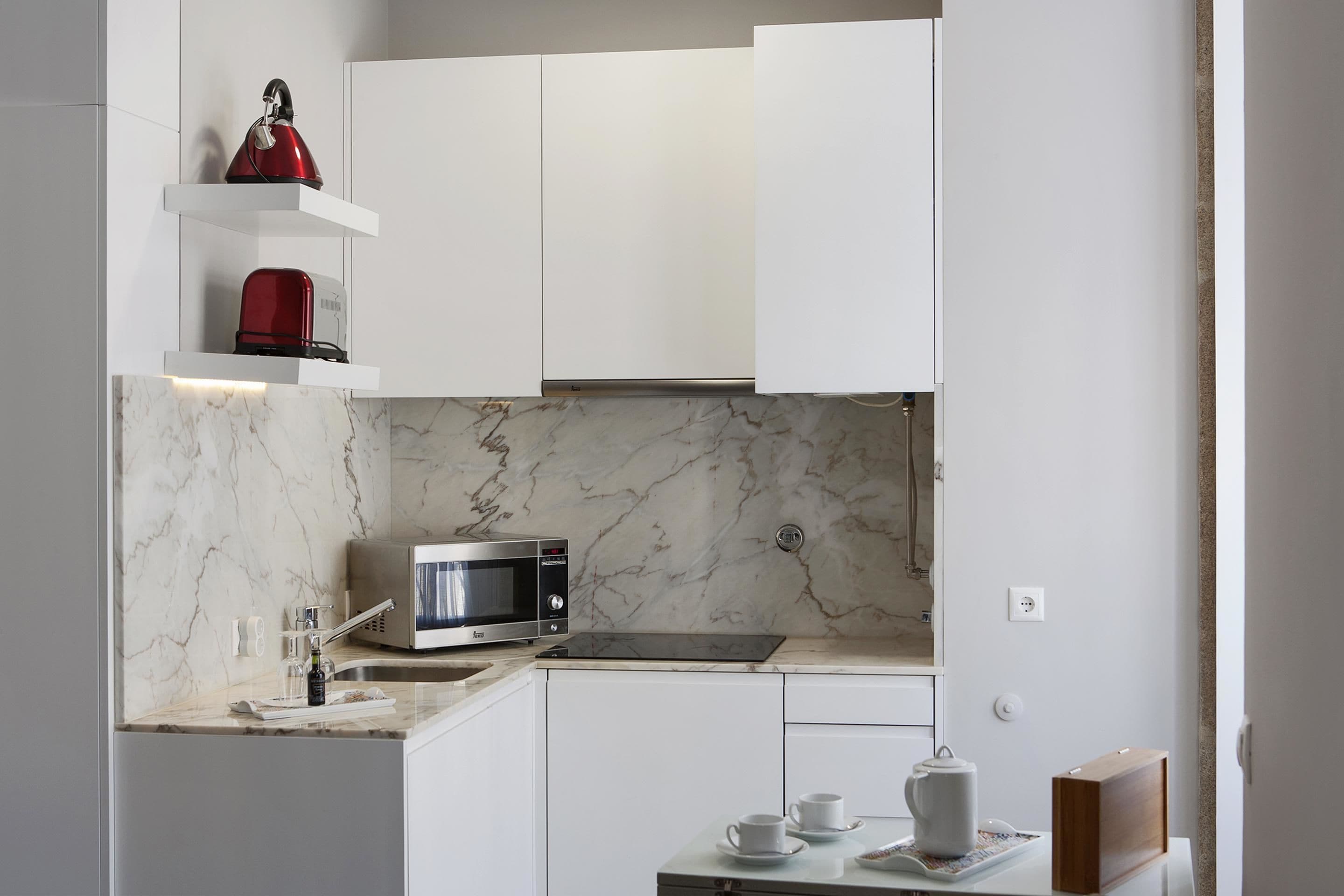 Une cuisine moderne avec des meubles blancs, un pare-feu en marbre, un four à micro-ondes et une bouilloire rouge posée sur une étagère.