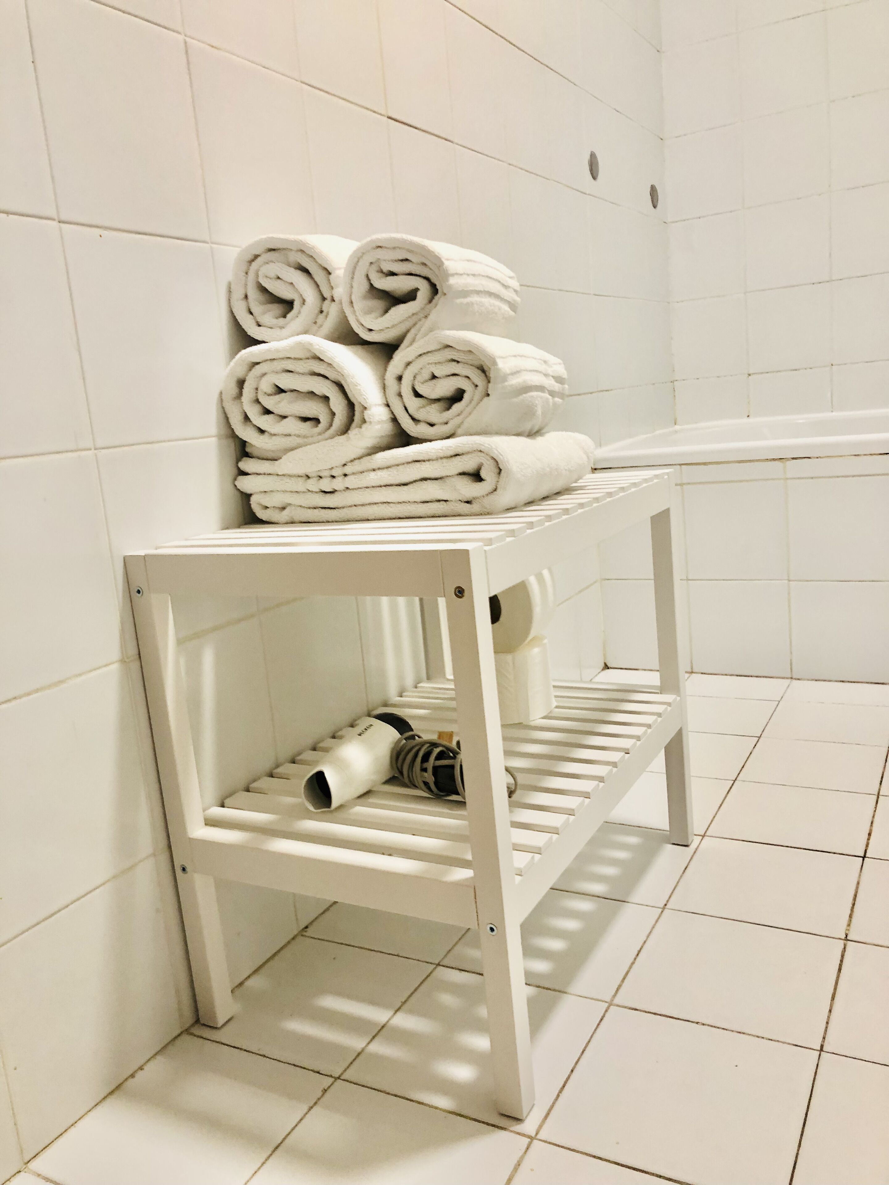 A white metal shelf with rolled white towels and a white towel dispenser in a bathroom with white tiled walls and floor.
