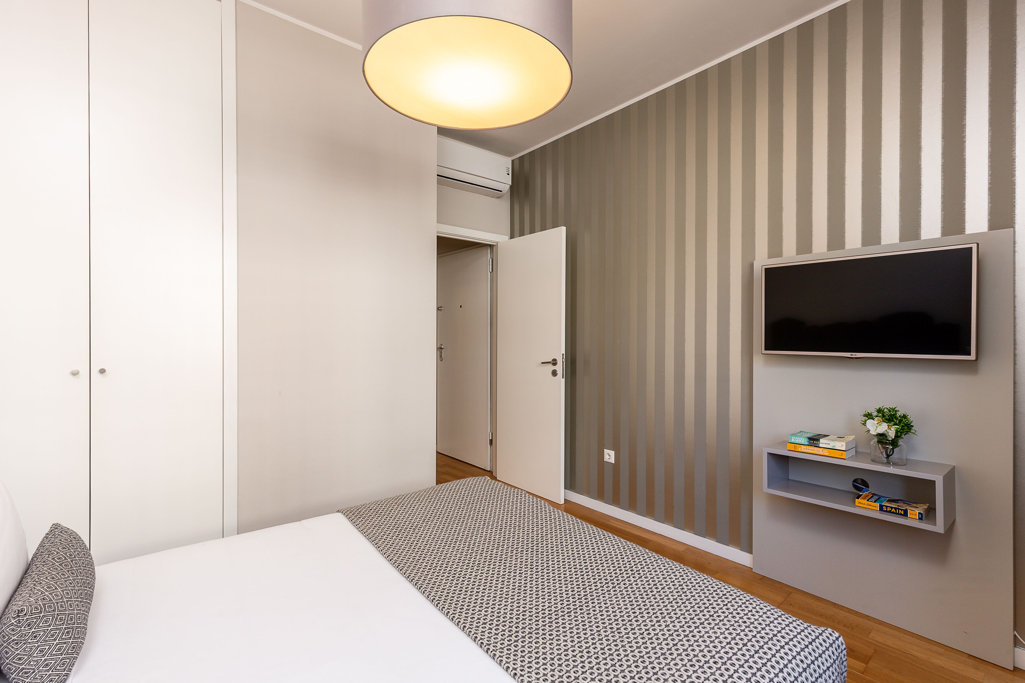 Ein Schlafzimmer mit Bett, Kleiderschrank, Fernseher und einem kleinen Regal mit Büchern und einer Pflanze.