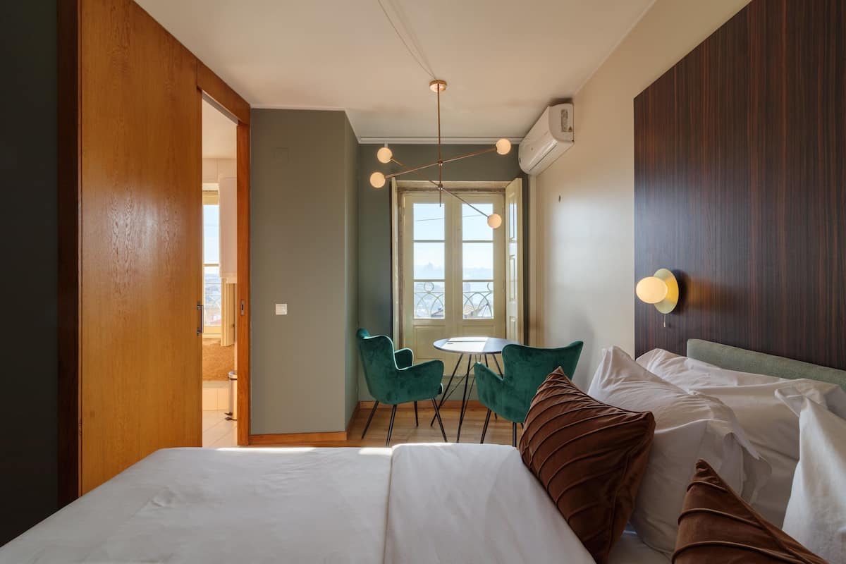 A hotel room with a bed, two green chairs, a small table, and a window with a view.