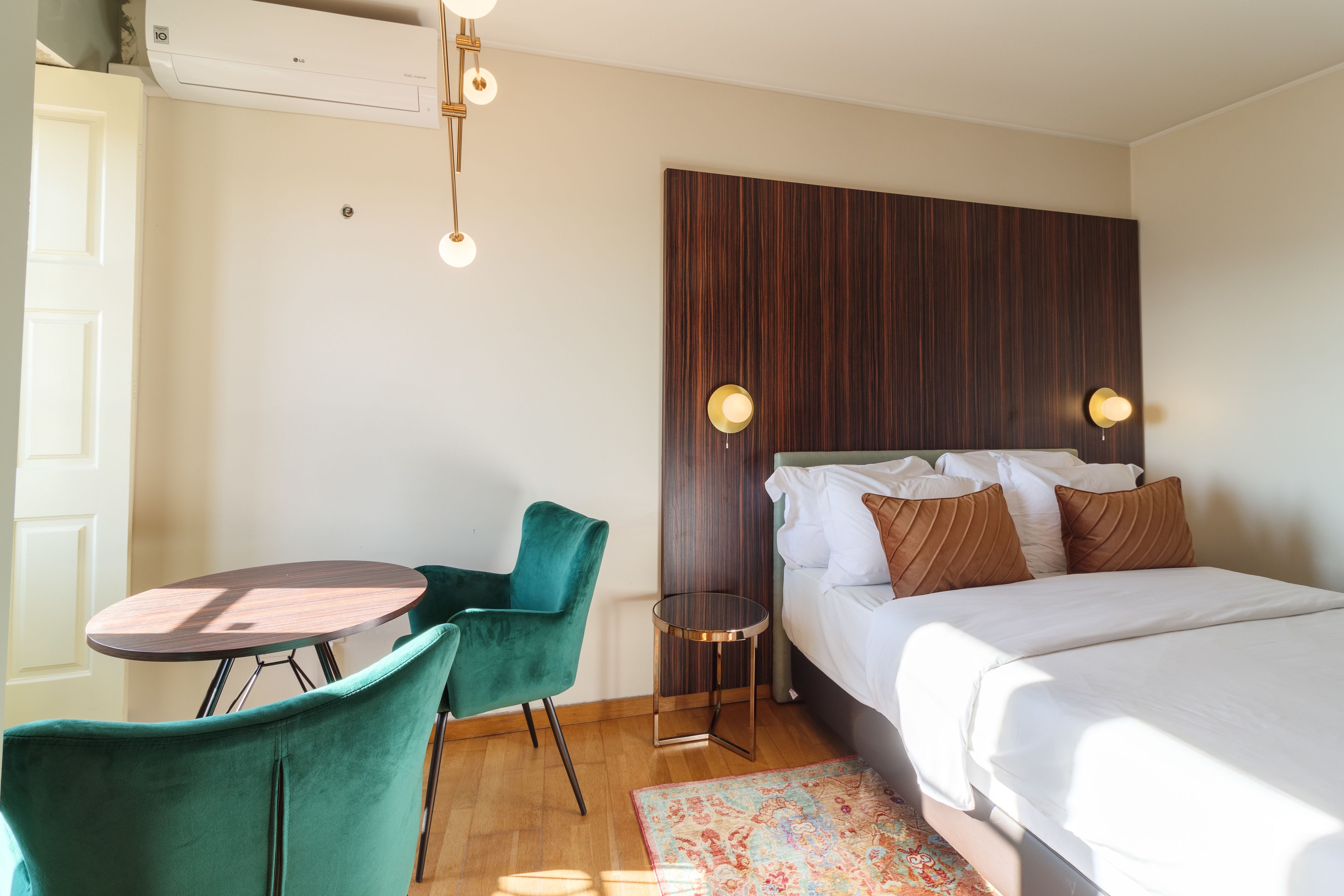A hotel room with a bed, a desk, a chair, a small table, and a wall-mounted light fixture.
