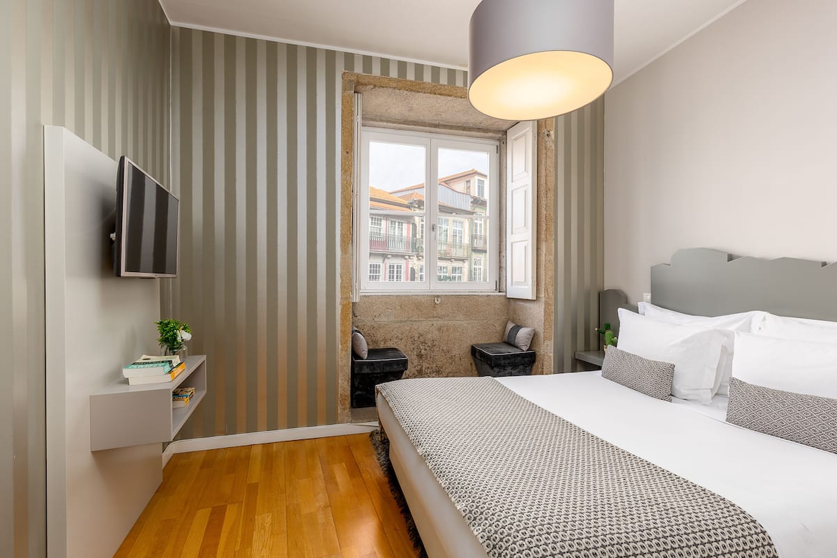 A modern hotel room with a large bed, a TV mounted on the wall, a small seating area, and a window with a view of buildings.