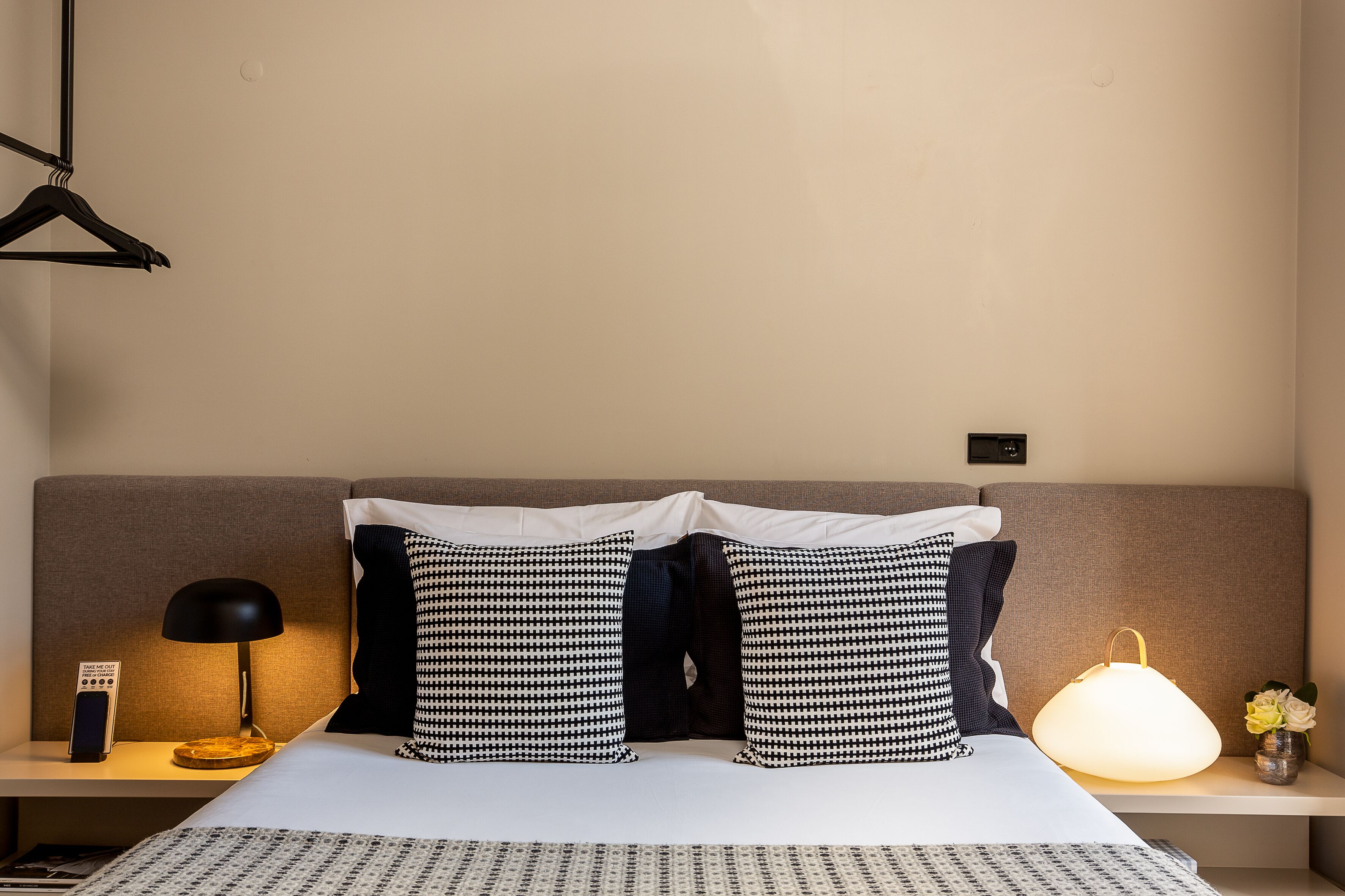 A neatly made bed with a white and patterned bedspread, two black and white patterned pillows, a black lamp on a bedside table, and a bedside table with a remote control and a small vase with flowers.