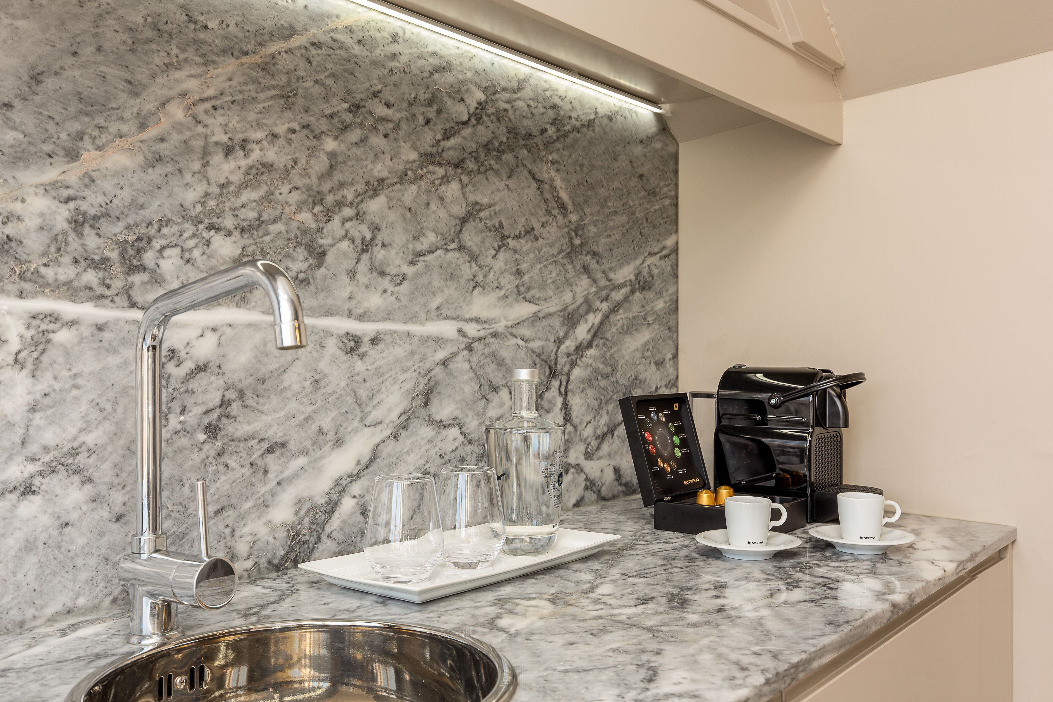 Una cocina con encimera de mármol, pileta de acero inoxidable, cafetera y una bandeja con vasos y una botella.