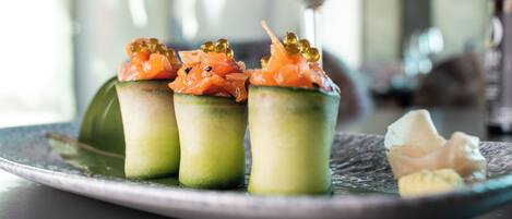 A plate with three sushi rolls topped with salmon and roe, a glass of white wine, and a small piece of white food on a dark plate.