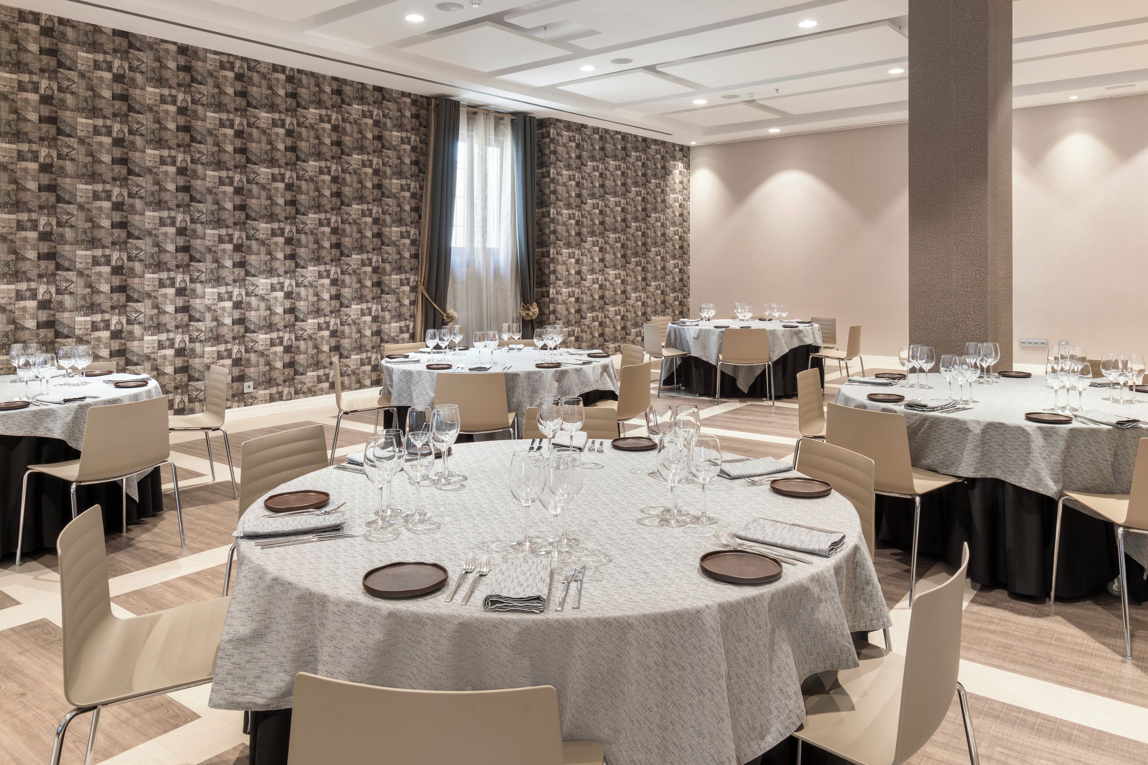 Un salón de conferencias con varias mesas redondas, cada una dispuesta con platos, cubiertos y copas de vino. La sala presenta un papel tapiz decorativo y un pilar grande.