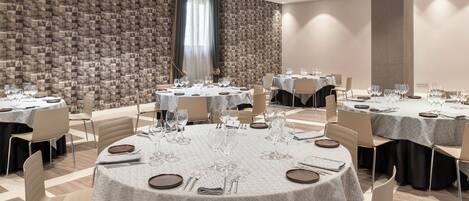 Un salón de conferencias con varias mesas redondas, cada una dispuesta con platos, cubiertos y copas de vino. La sala presenta un papel tapiz decorativo y un pilar grande.