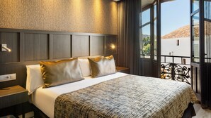A neatly made bed with a patterned bedspread, a bedside table with a lamp, and a view of a building through a window.