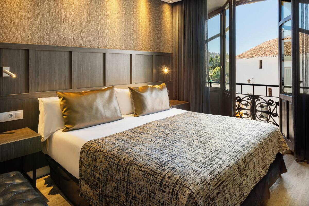 A neatly made bed with a patterned bedspread, a bedside table with a lamp, and a view of a building through a window.