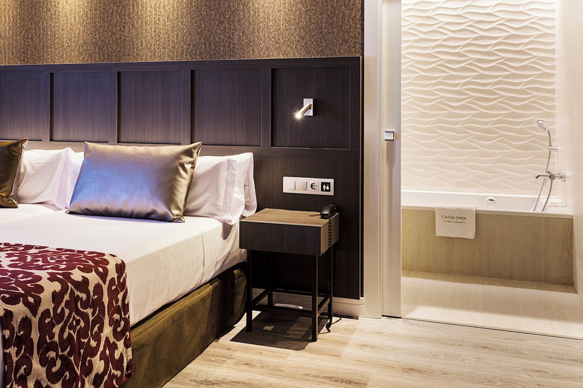 Una habitación de hotel con una cama, mesita de noche y un baño con bañera.