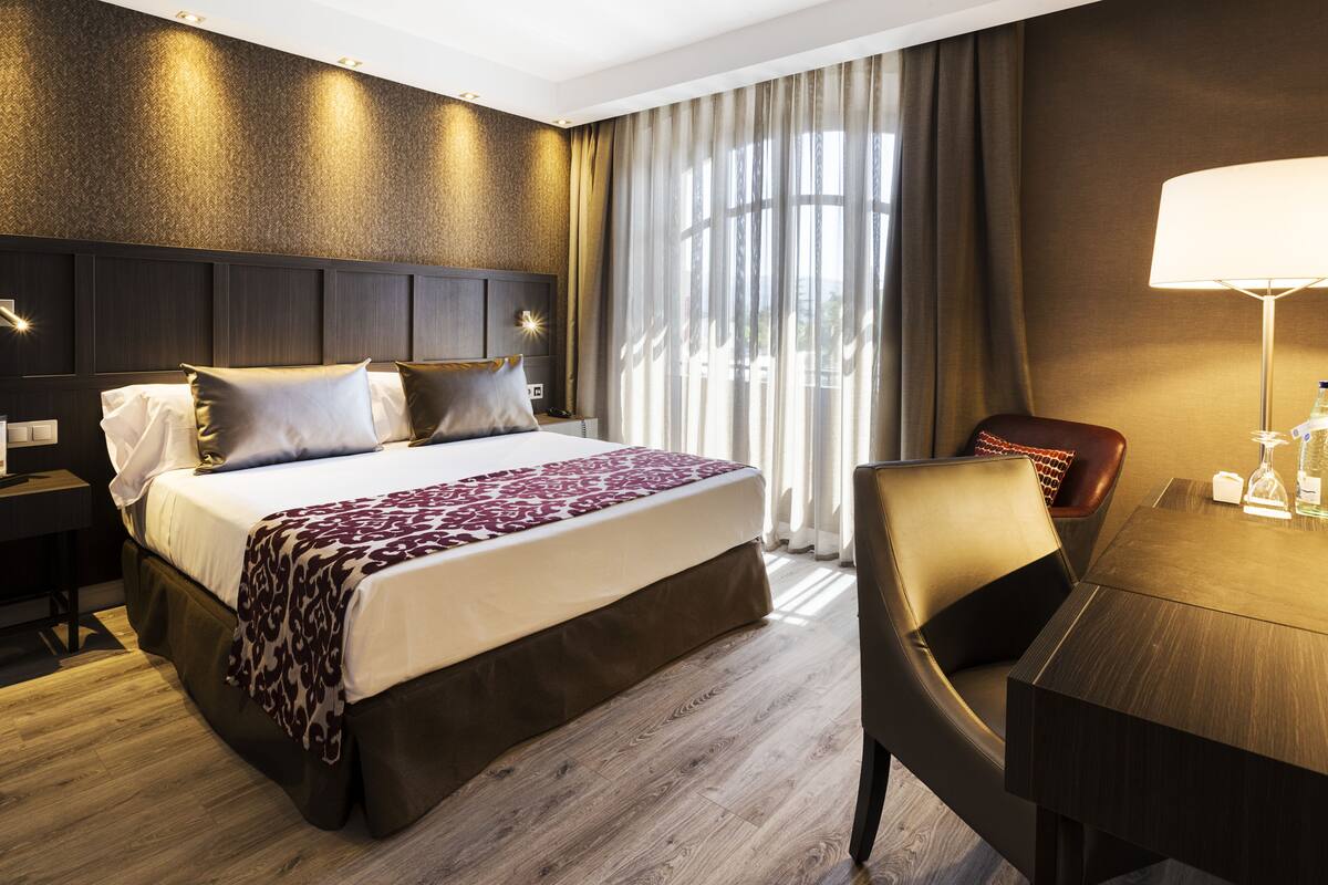 A hotel room with a large bed, a desk, a chair, a lamp, and a view of the outdoors.
