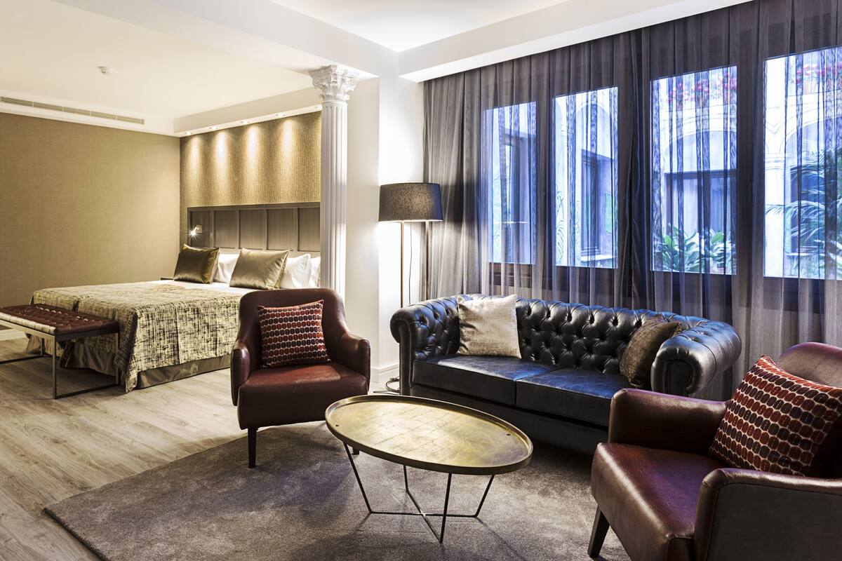 A hotel room with a bed, a sofa, two armchairs, a coffee table, and large windows with curtains.