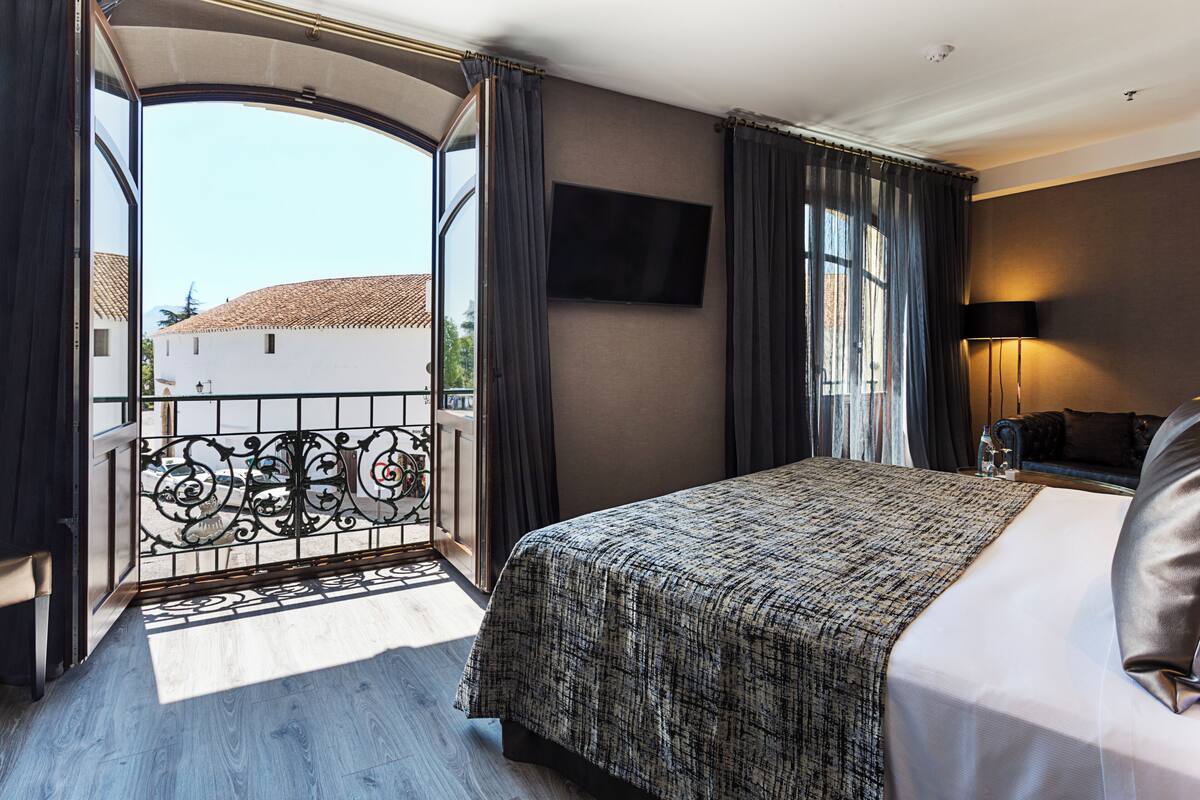 A hotel room with a bed, a TV, a balcony with a view of buildings, and a door leading outside.