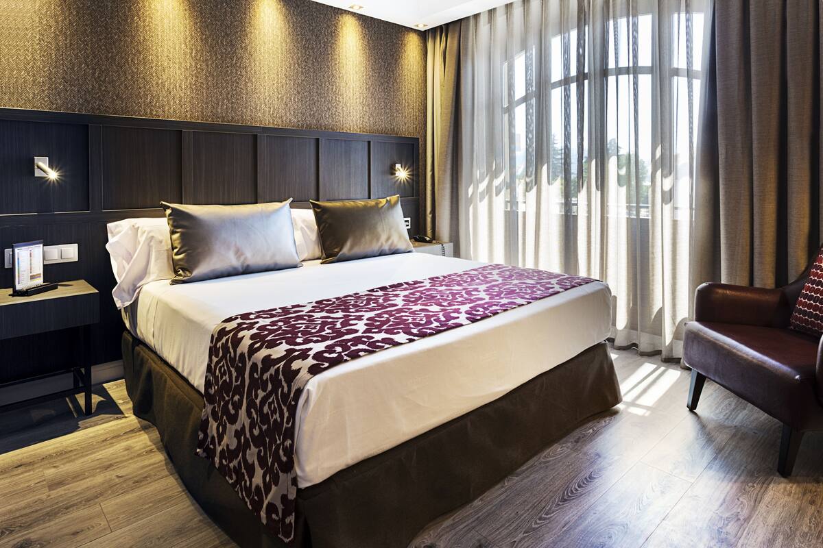 A hotel room with a large bed, a nightstand, a chair, and a window with curtains.