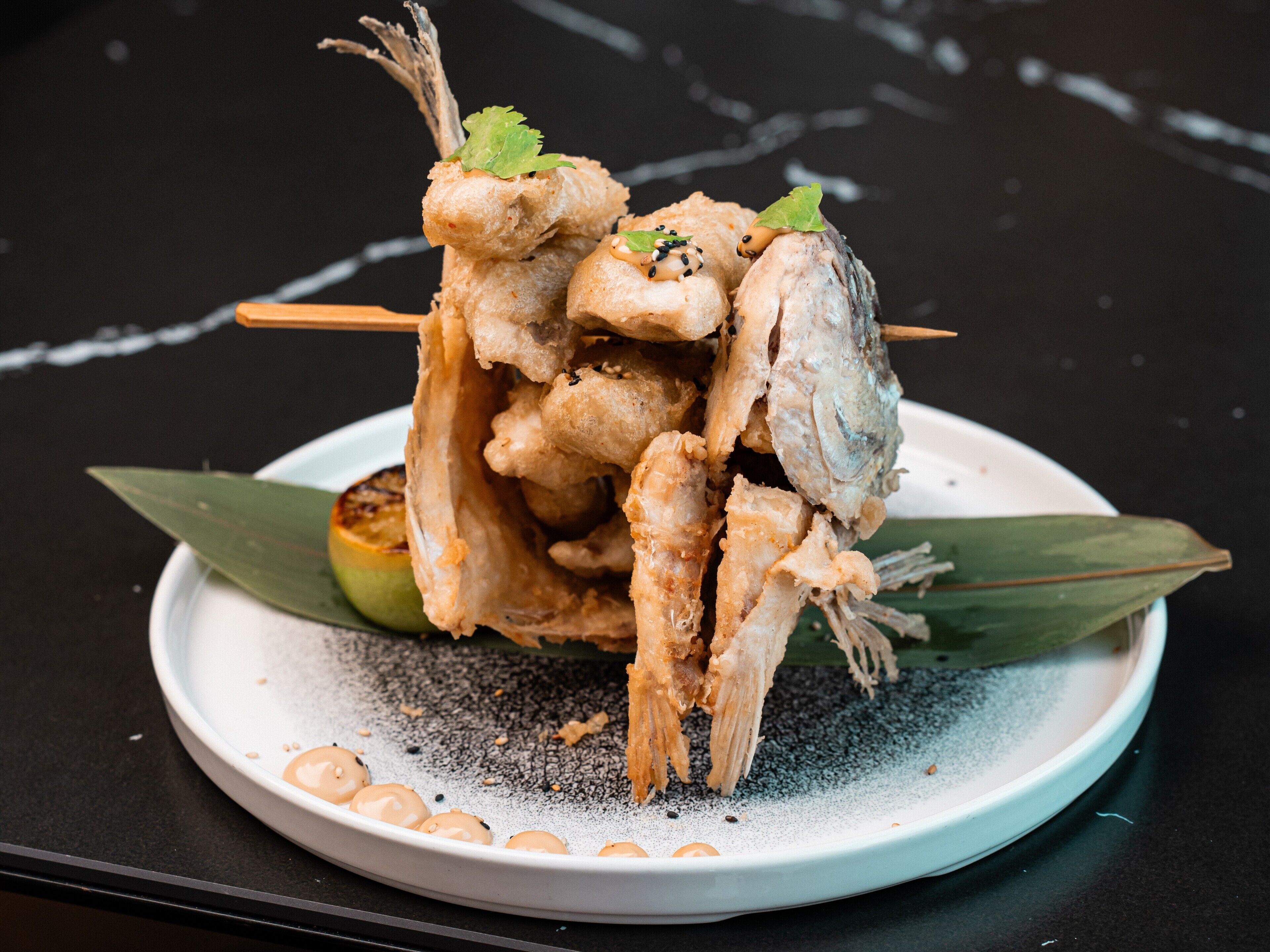 A plate of fried fish with a garnish of black sesame seeds and a green leaf, served on a white plate with a black speckled pattern.