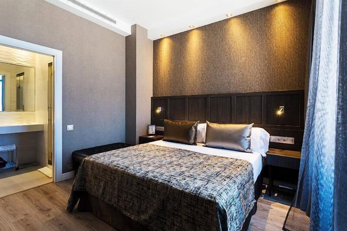 A modern hotel room with a large bed, a headboard with built-in lighting, a bedside table with a lamp, and a wardrobe.