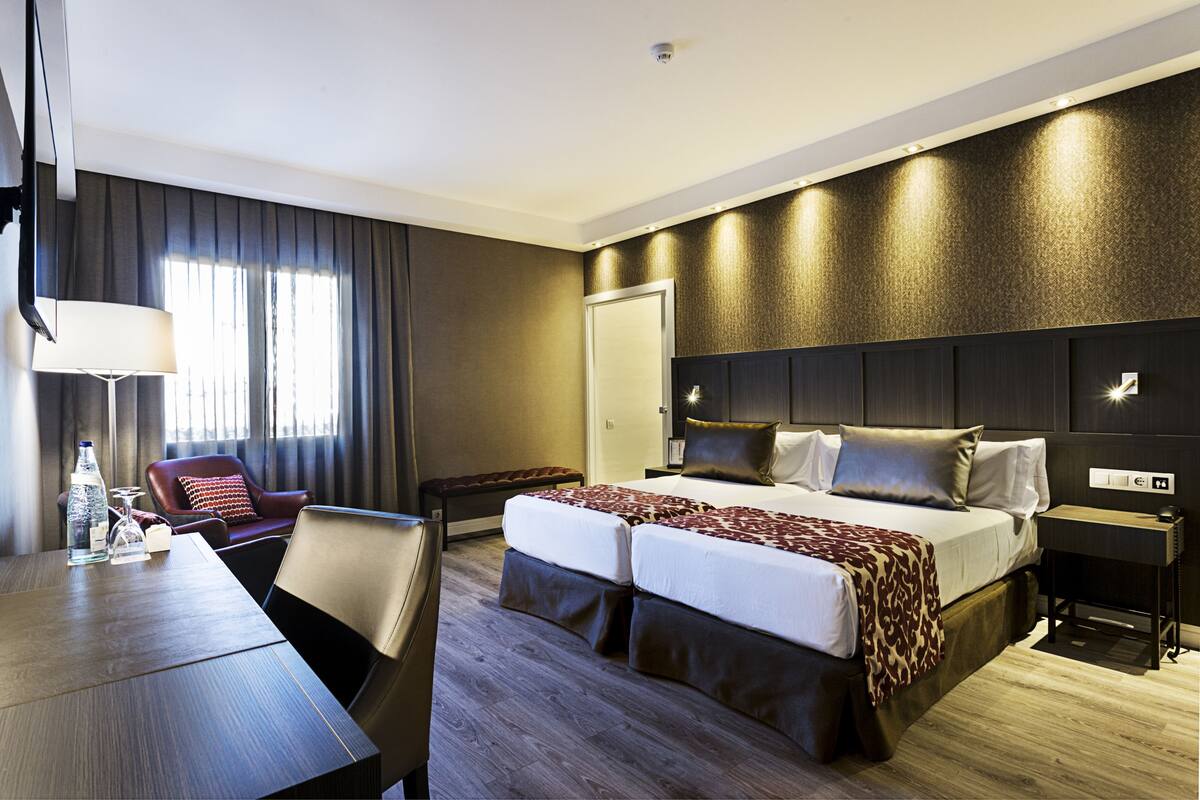 A hotel room with a large bed, a desk with a lamp, a chair, and a window with curtains.