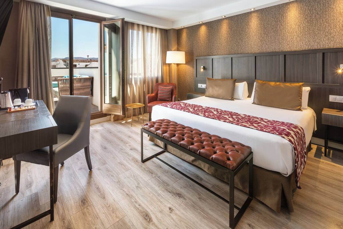 A hotel room with a large bed, a desk with a chair, a bench, a lamp, and a view of the city.