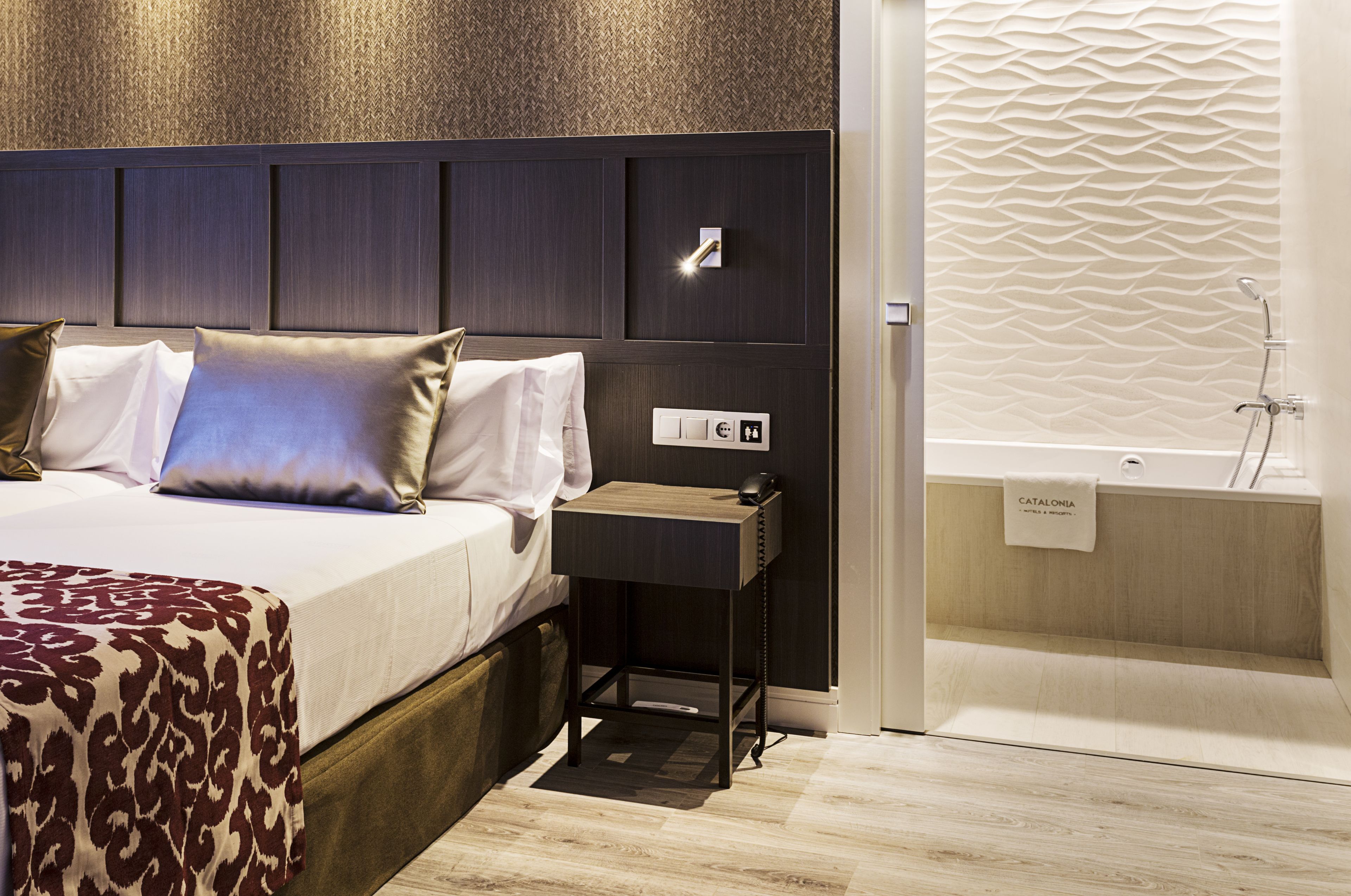 A modern hotel room with a bed, bedside table, and a bathroom with a bathtub.