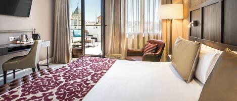 A hotel room with a large bed, a desk with a chair, a TV, and a view of the city through the window.