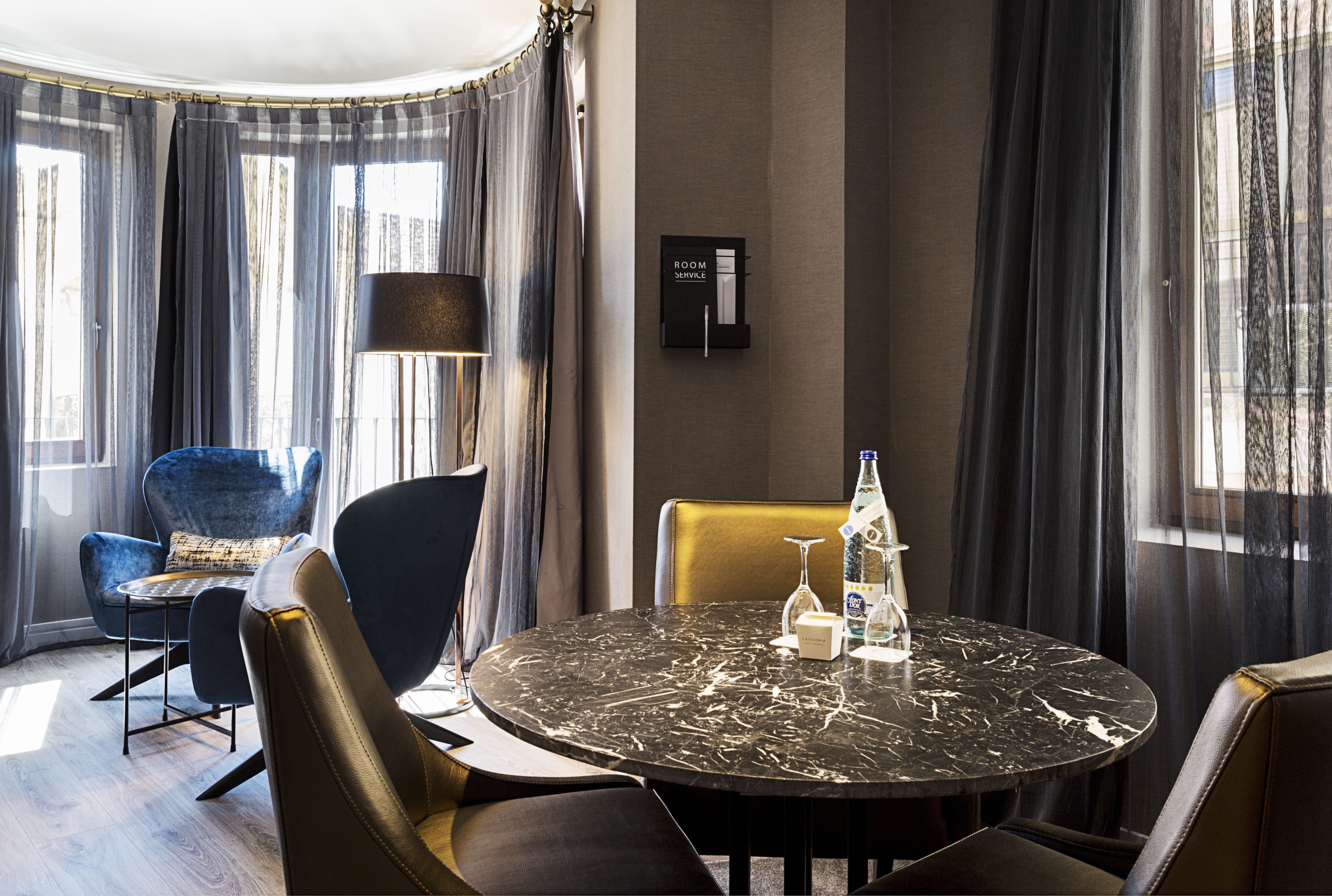 A modern hotel room with a round marble table, leather chairs, a standing lamp, and a view of the city through large windows with sheer curtains.