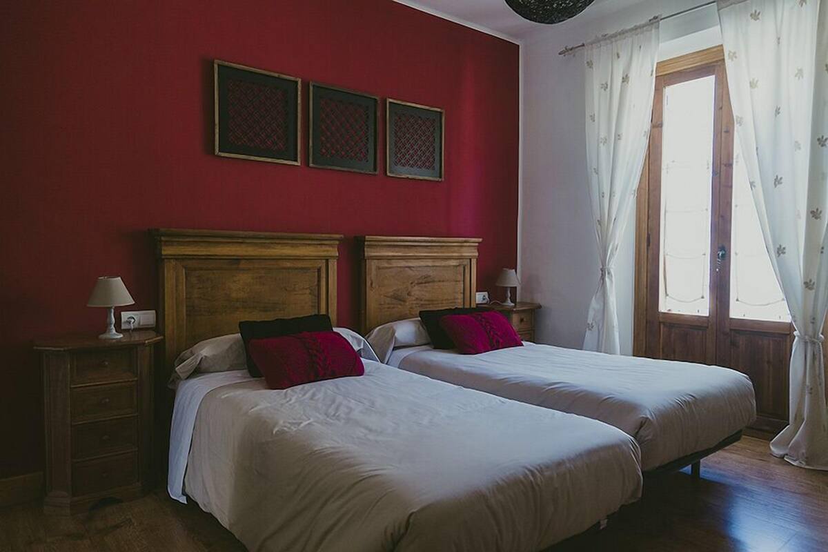 A room with two beds, wooden headboards, a red wall, and a window with white curtains.