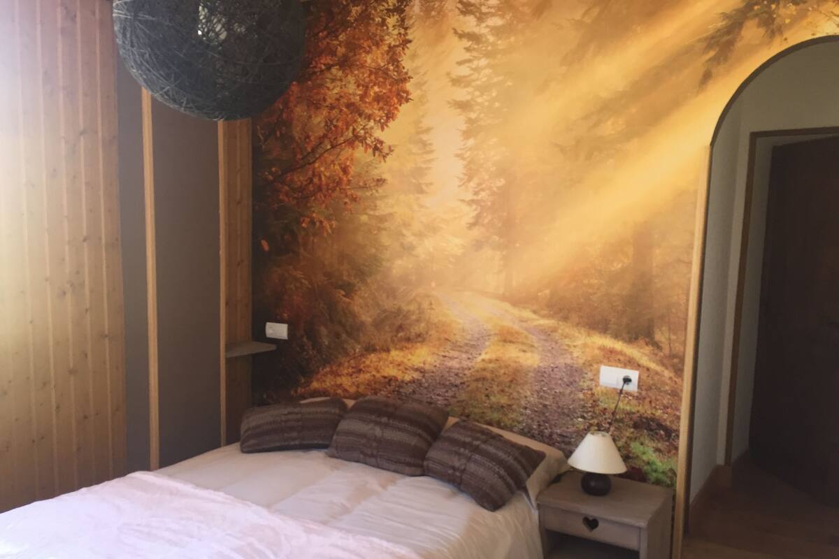 A bedroom with a large bed, a nightstand with a lamp, and a wall mural of a forest scene.