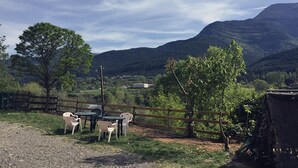 An outdoor seating area with tables and chairs, surrounded by a wooden fence and trees, overlooking a valley with mountains in the background. - Leyendas del Pirineo (Fiscal)