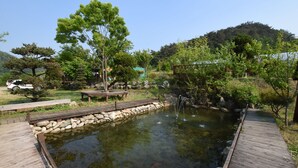 A wooden deck leading to a pond with a small waterfall, surrounded by trees and a stone wall. - Ecopia Pension (Pyeongchang)