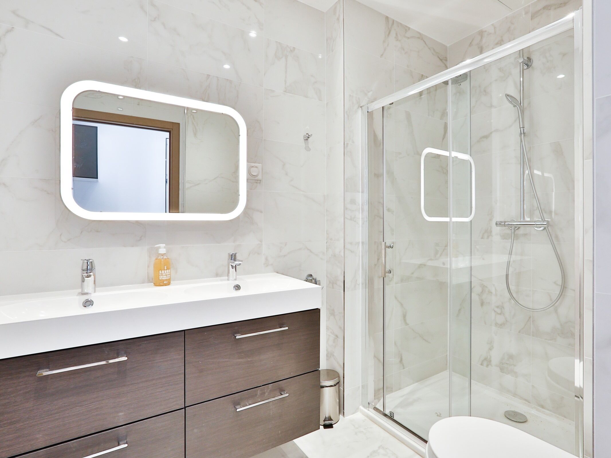 Een moderne badkamer met een douchecabine van glas, een wit wastafelmeubel met een marmeren aanrechtblad en een grote spiegel boven de wastafel.