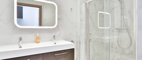 Een moderne badkamer met een douchecabine van glas, een wit wastafelmeubel met een marmeren aanrechtblad en een grote spiegel boven de wastafel.