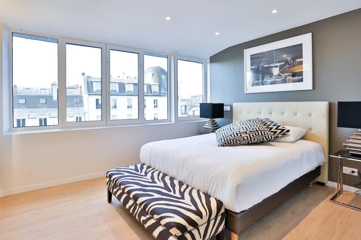 A modern bedroom with a large bed, a bench, and a view of buildings through large windows.