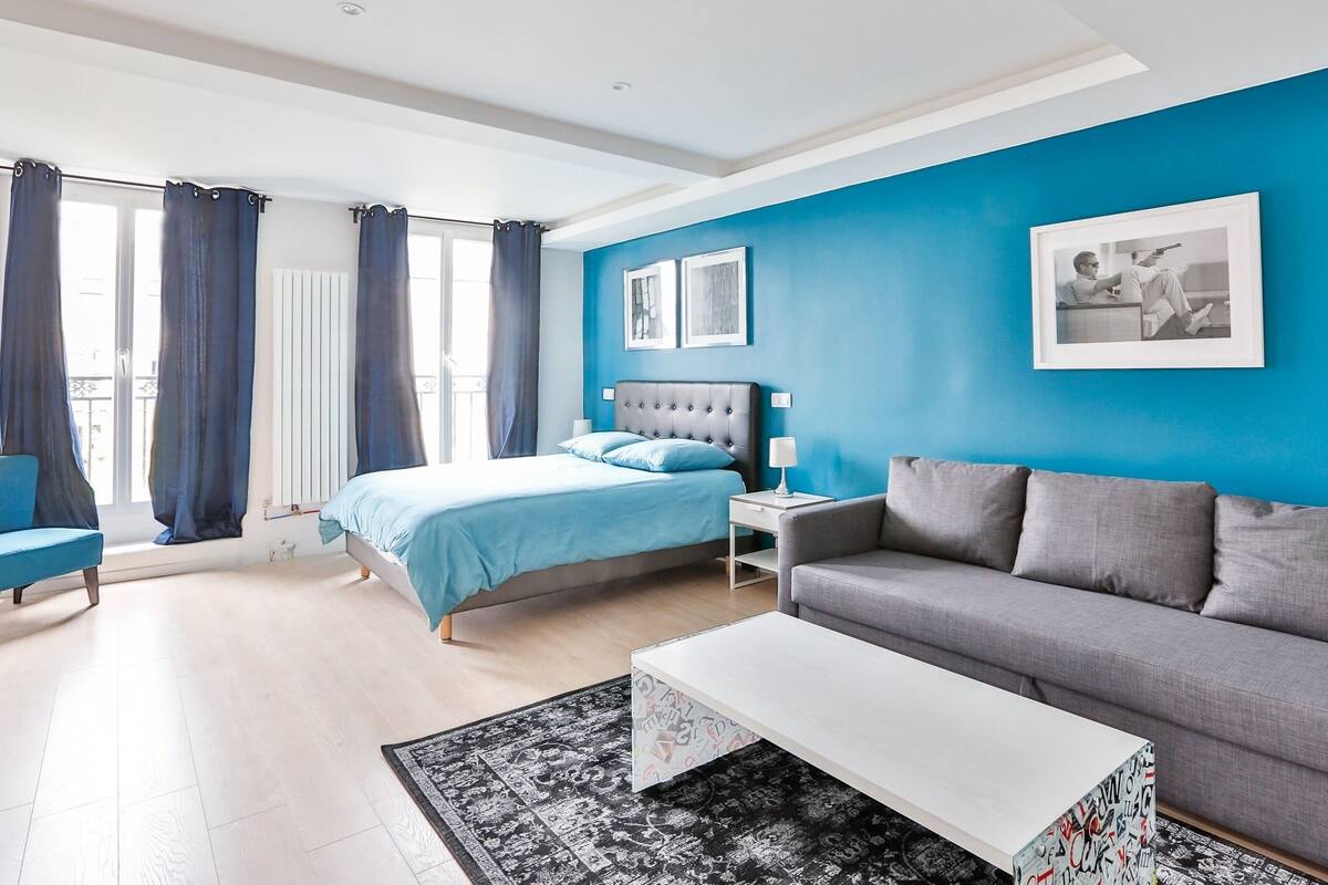 A modern bedroom with a bed, a grey sofa, a white coffee table, and a blue accent wall.