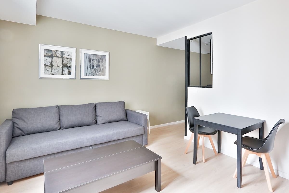 A modern living room with a grey sofa, a small table, and two chairs. There are two framed pictures on the wall.