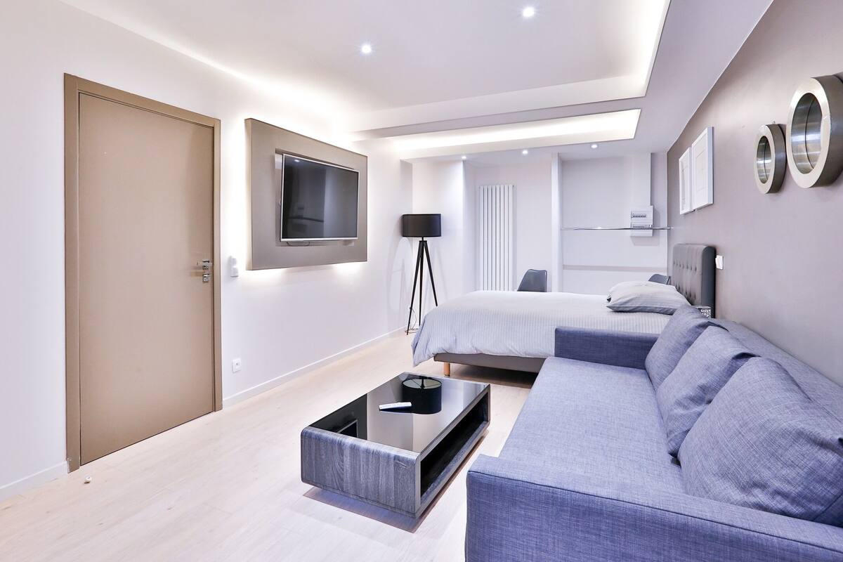A modern hotel room with a large bed, a sofa, a TV mounted on the wall, and a desk with a lamp.