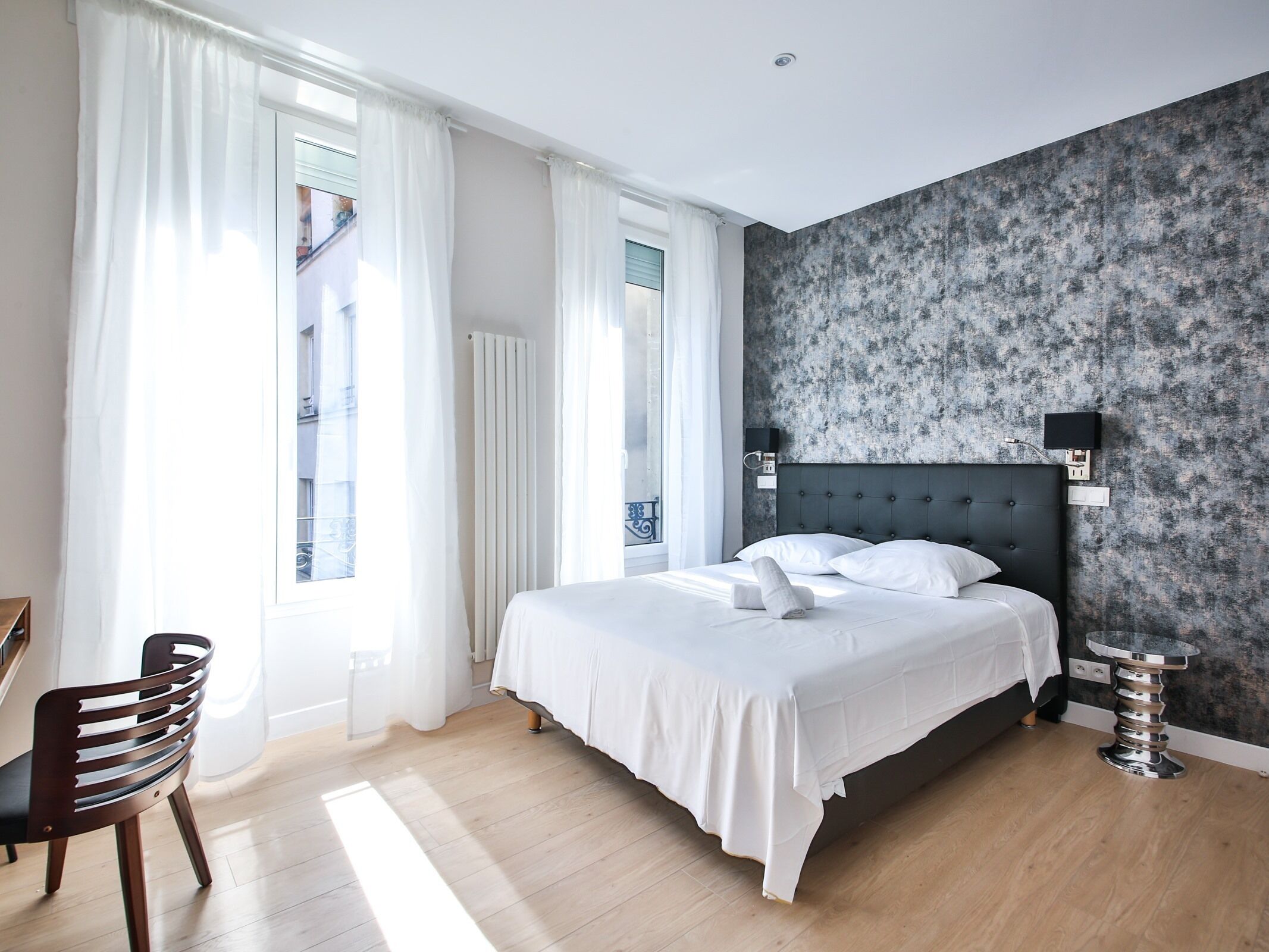 Un dormitorio moderno con una cama grande, una silla, una mesita redonda y vistas al exterior por la ventana.