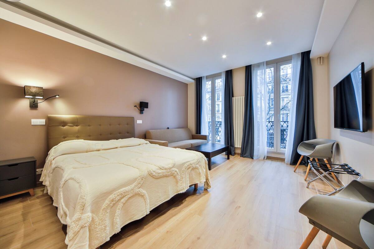 A modern bedroom with a large bed, a sofa, a TV, and a window with curtains.