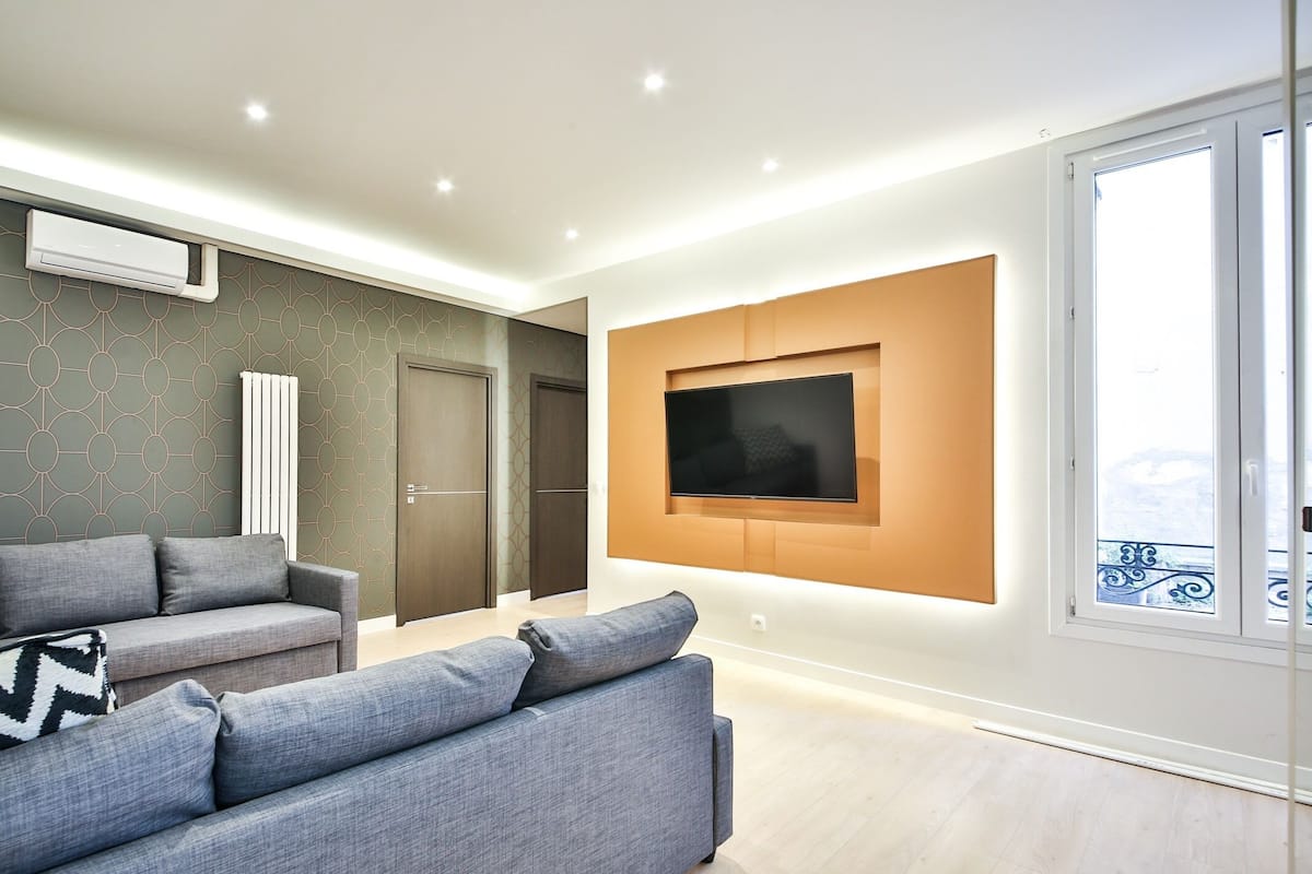 A modern living room with a grey sofa, a flat-screen TV mounted on the wall, and a large window providing a view of the outside.