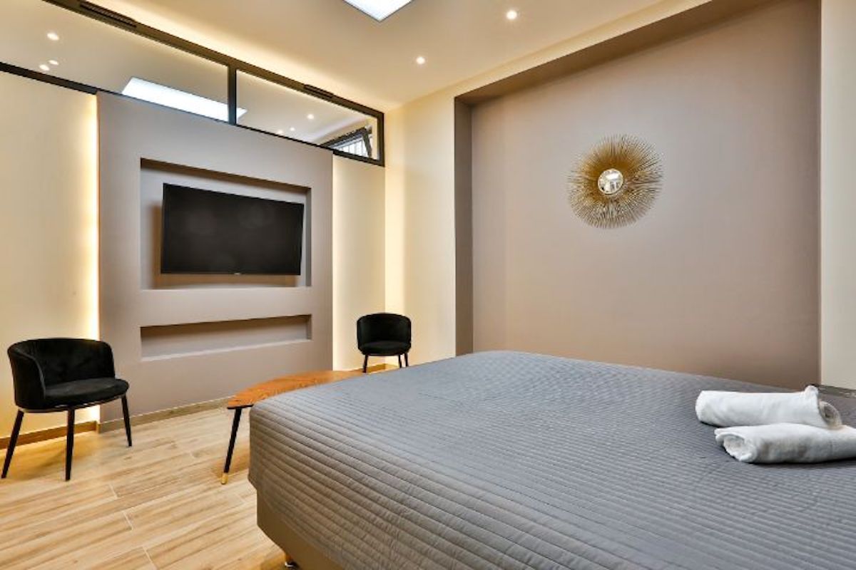 Una habitación de hotel moderna con una cama grande, un televisor de pantalla plana montado en la pared, dos sillas negras y un adorno decorativo en la pared.