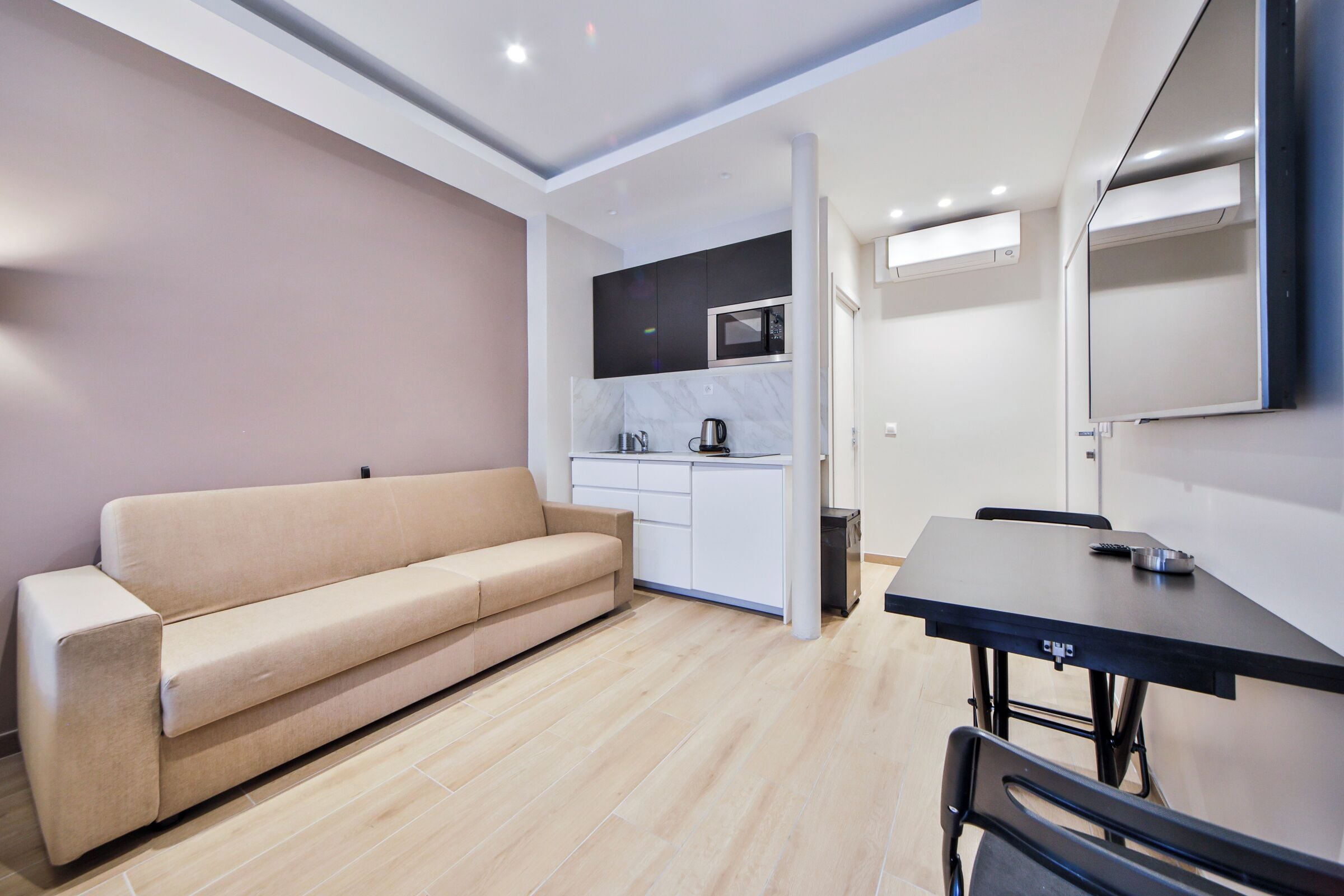 A modern living room with a beige sofa, a dining table with chairs, and a kitchen area with a microwave and cabinets.