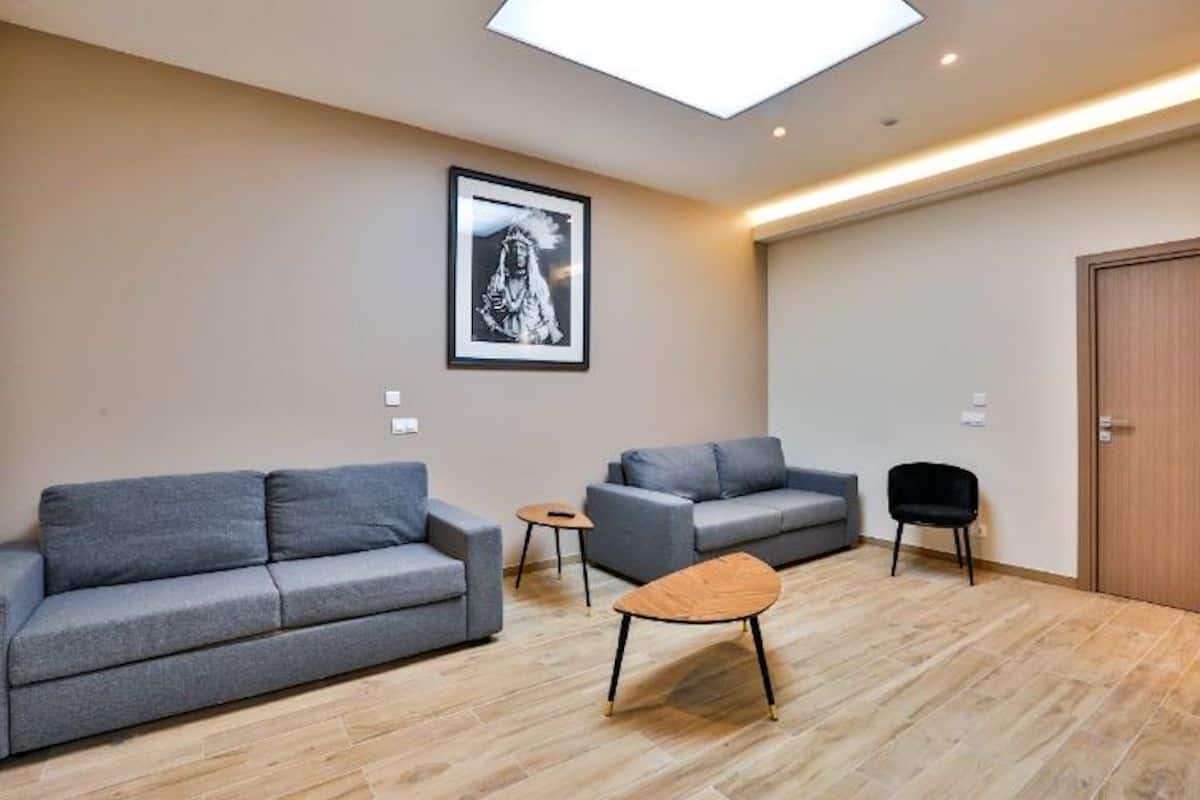 A modern living room with two grey sofas, a small round table, and a wooden coffee table. A framed picture hangs on the wall, and a door is visible in the background.